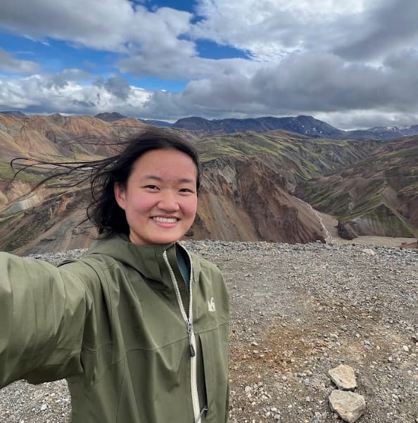 Maggie smiling in a selfie taken on a rocky summit, with colorful mountain ridges and cloudy sky behind her